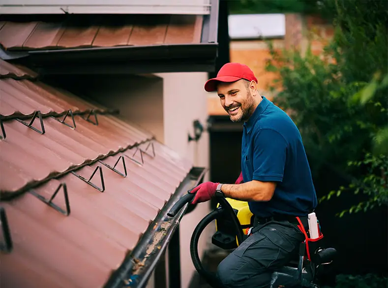 Happy maintenance worker cleaning gutters on roof with blower.
